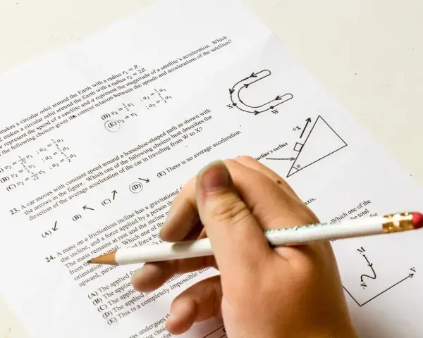 Ręka z ołówkiem rozwiązuje test fizyczny. Pytanie 22 dotyczy satelitów, a pytanie 23 ruchu po torze w kształcie podkowy. Od ilu procent jest 5?