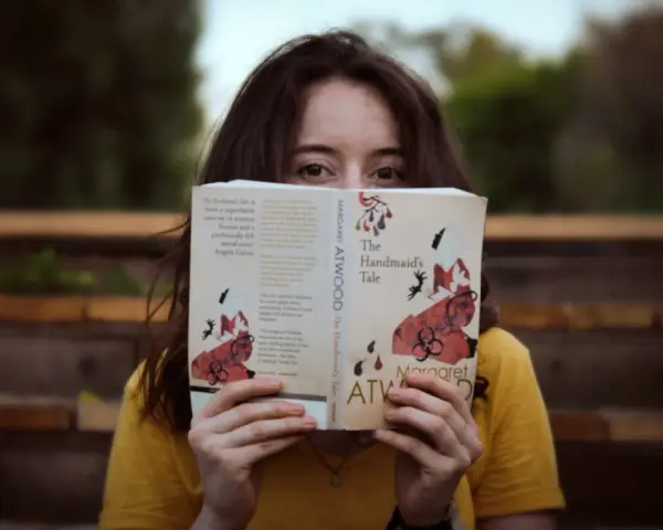 Młoda kobieta w żółtej bluzce czyta "Opowieść podręcznej" Margaret Atwood. Jej oczy błyszczą, gdy zagłębia się w świat literatury, co jest idealnym przykładem pasji do filologii.