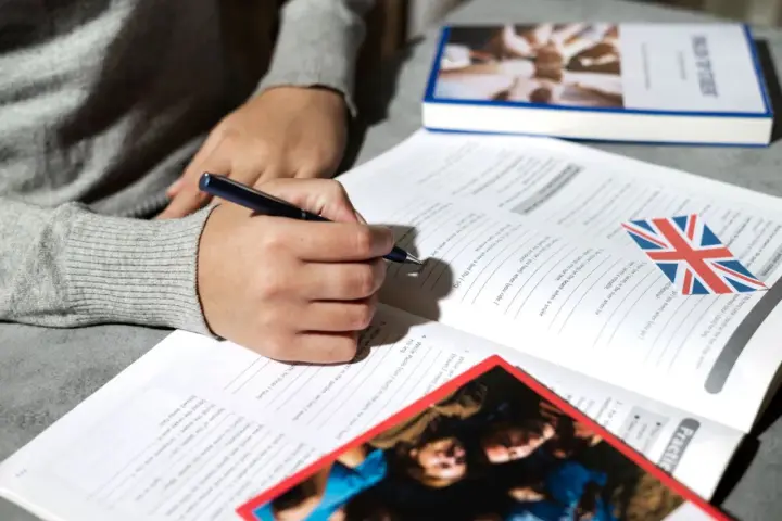 Osoba pisze długopisem w zeszycie z ćwiczeniami z języka angielskiego. Obok leży książka i flaga Wielkiej Brytanii. To pomoc w nauce, jak klauzula CV po angielsku.