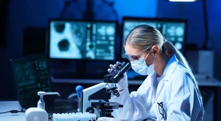 Diagnosta laboratoryjny w okularach i masce analizuje próbki pod mikroskopem. Na ekranach komputera widać wyniki badań.