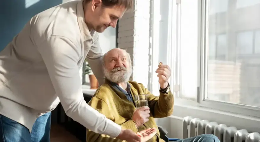 Mężczyzna w średnim wieku podaje starszemu mężczyźnie w wózku inwalidzkim tabletkę i szklankę wody.
