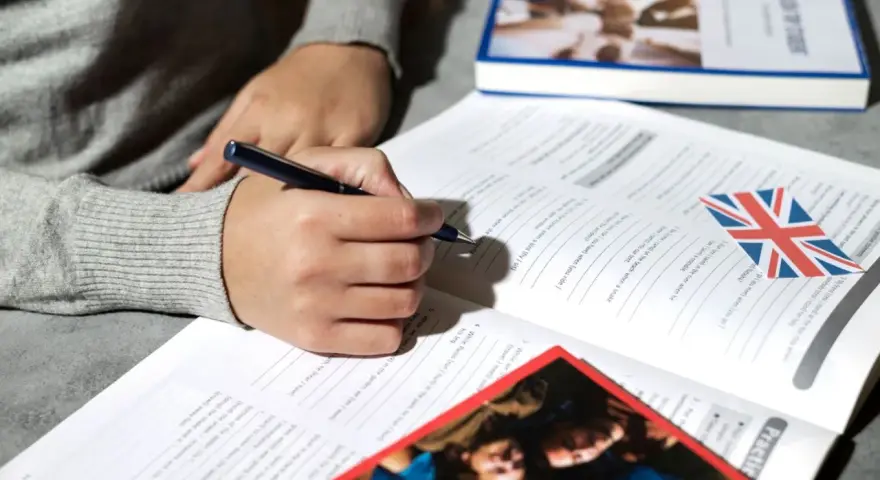 Osoba pisze długopisem w zeszycie z ćwiczeniami z języka angielskiego. Obok leży książka i flaga Wielkiej Brytanii. To pomoc w nauce, jak klauzula CV po angielsku.