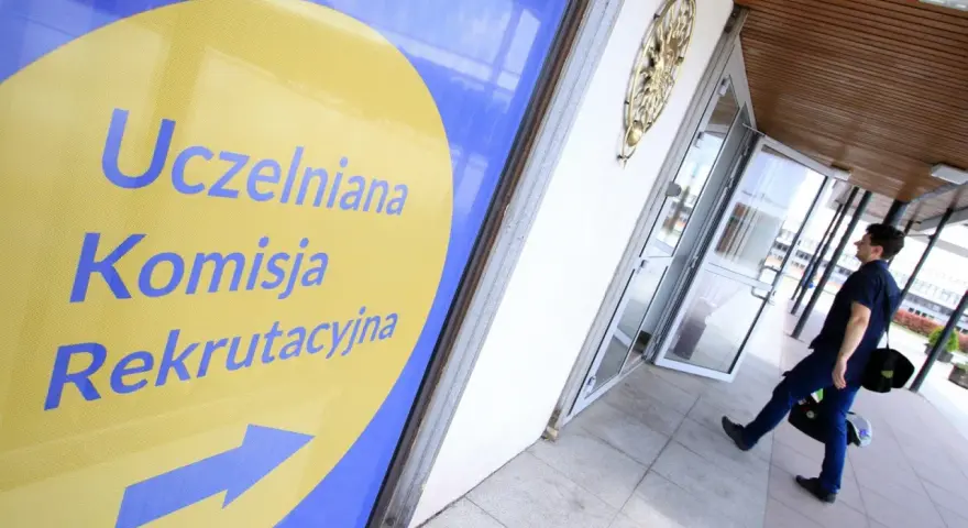 Znak "Uczelniana Komisja Rekrutacyjna" wskazuje drogę do budynku, gdzie trwa rekrutacja na studia. Mężczyzna z torbą idzie w kierunku drzwi.