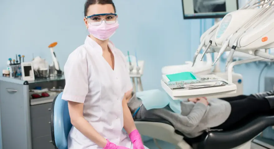 Asystentka stomatologiczna w masce i okularach ochronnych siedzi w gabinecie.