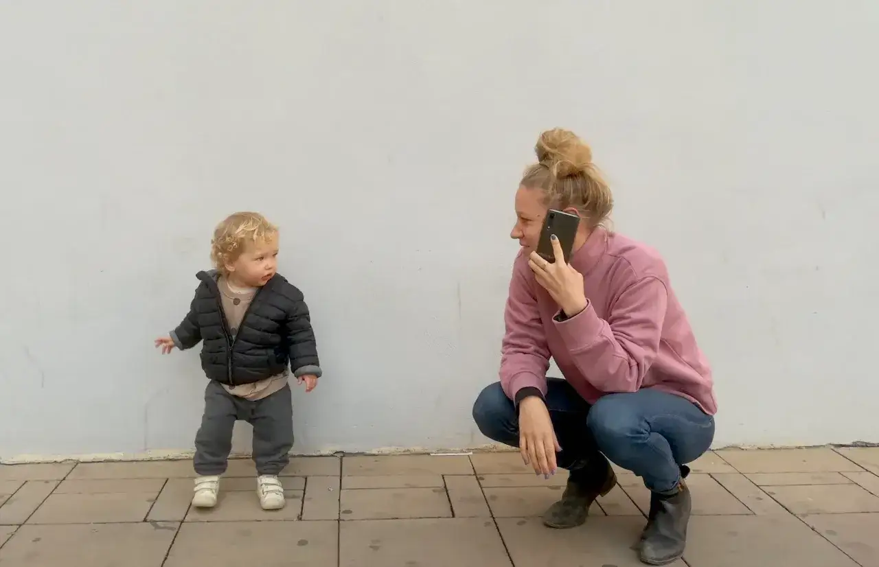 Mama rozmawia przez telefon, podczas gdy jej dziecko bawi się obok. To obraz codzienności, w której rodzicielstwo i wsparcie, jak rodzinny kapitał opiekuńczy, przeplatają się z obowiązkami.