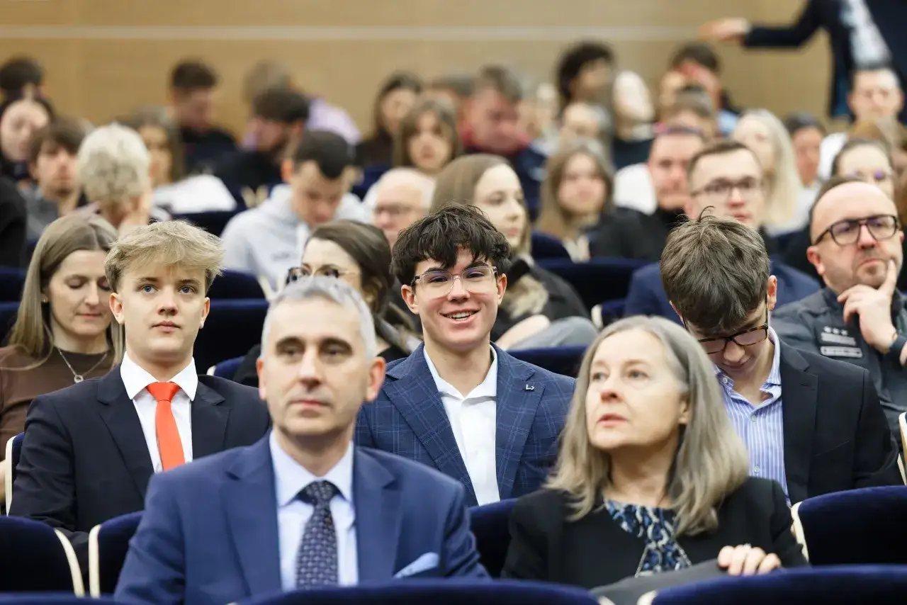 Uczestnicy konferencji z politologii, wśród nich młodzi ludzie w garniturach i eleganckie kobiety, słuchają wykładu.