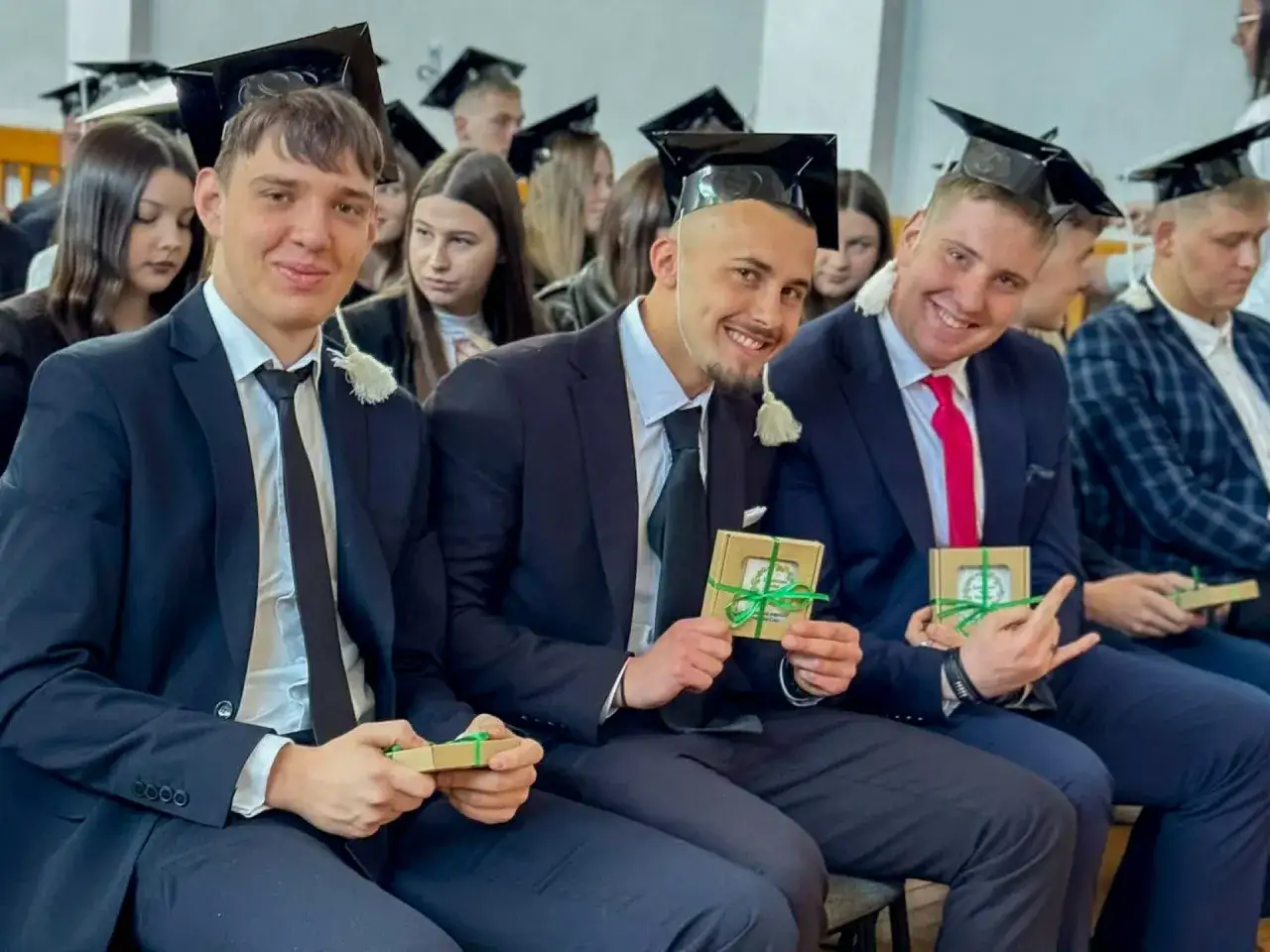 Uśmiechnięci maturzyści w togach i biretach świętują zakończenie roku szkolnego, trzymając w rękach drobne upominki.