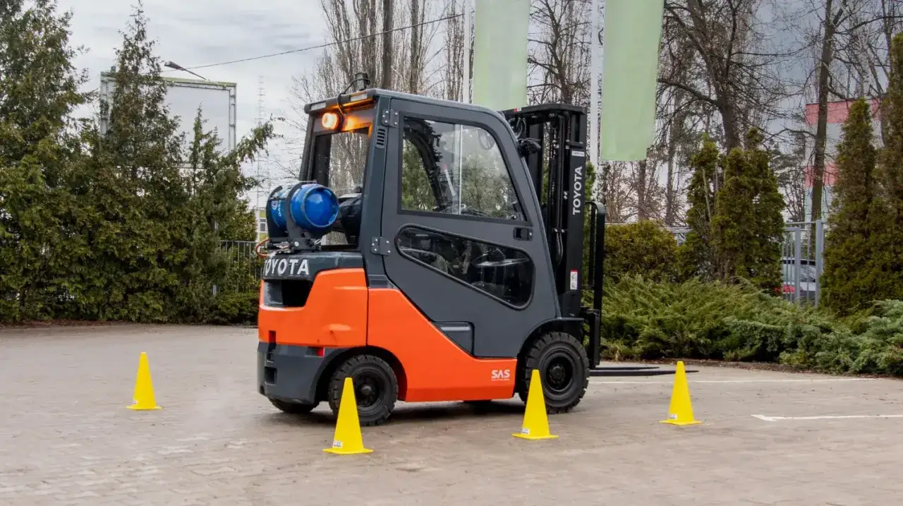 Pomarańczowo-szary wózek widłowy Toyota z butlą gazową, gotowy do pracy. Operator wózka widłowego ćwiczy manewrowanie między żółtymi pachołkami.