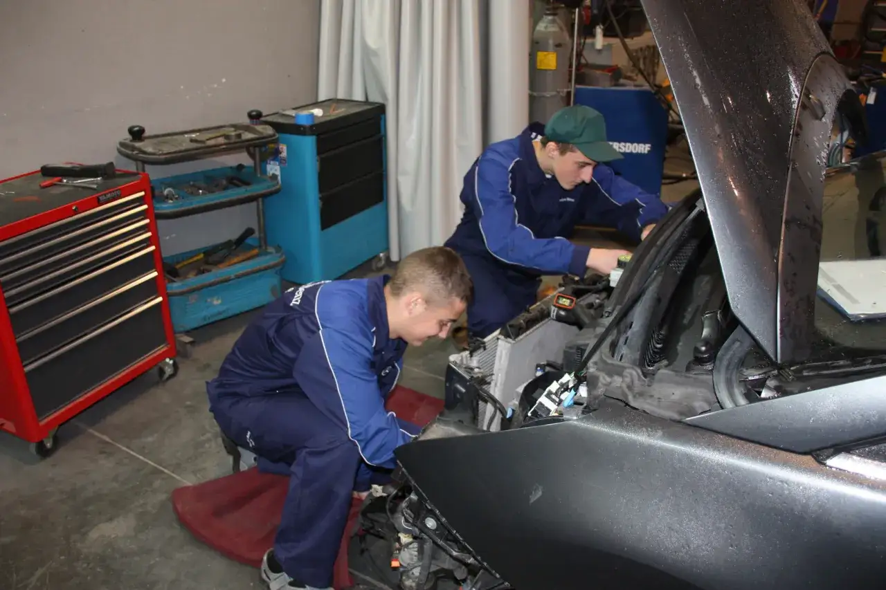 Dwóch młodych mechaników w niebieskich kombinezonach odbywa praktyki zawodowe, pracując przy samochodzie.