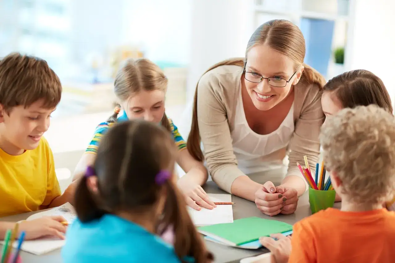 Uśmiechnięta nauczycielka wspiera dzieci w nauce. Widać zeszyty, kredki i zaangażowanie w pedagogikę.