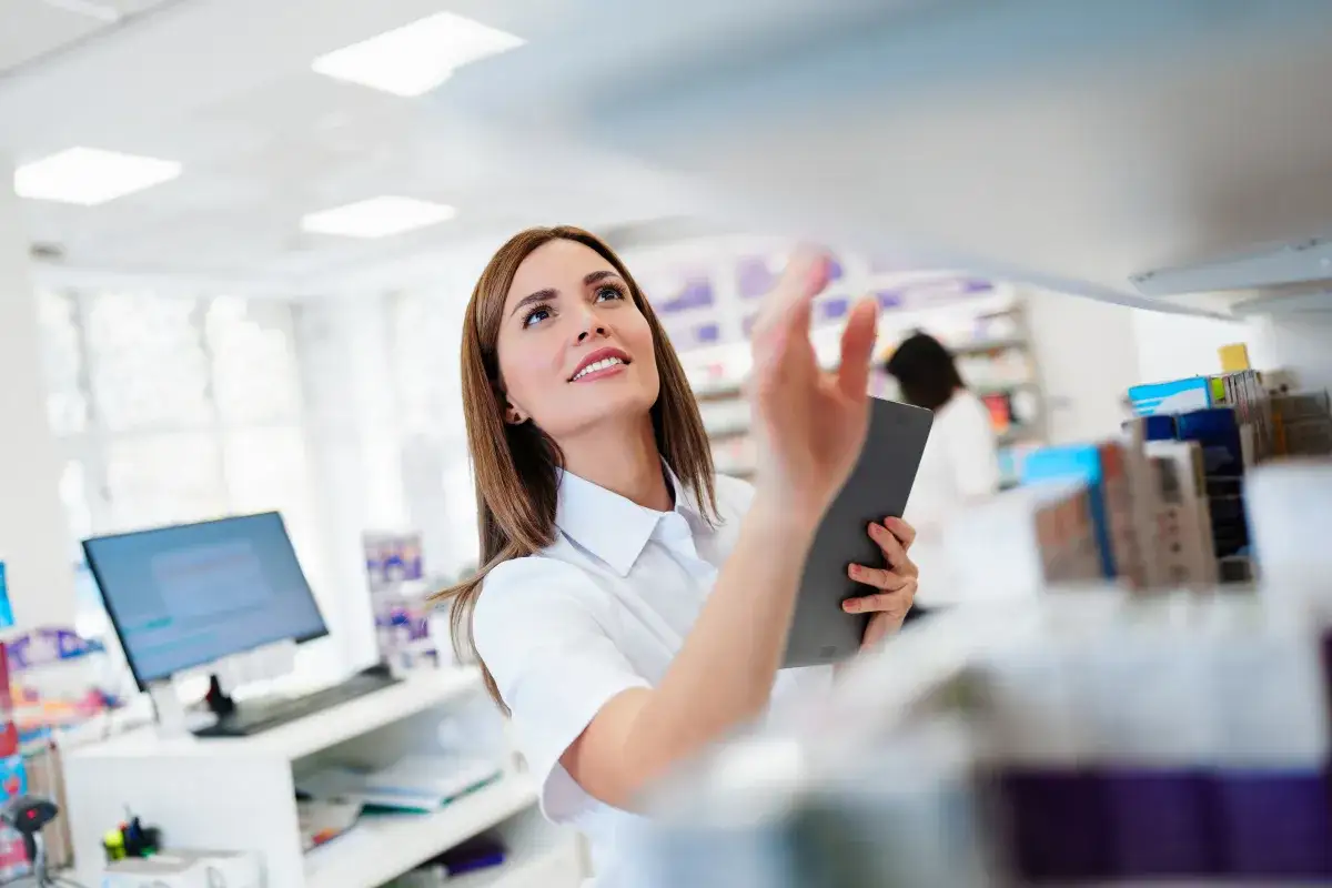Uśmiechnięta technik farmacji z tabletem w ręku sprawdza zapasy na półce w aptece.