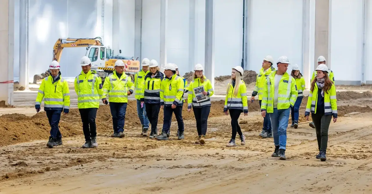 Grupa inżynierów w kamizelkach odblaskowych i kaskach zwiedza plac budowy. W tle koparka.