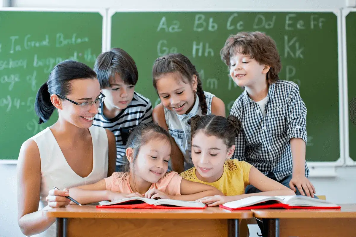 Nauczycielka z dziećmi przy książkach. W tle tablica z literami. To przykład pedagogiki przedszkolnej i wczesnoszkolnej.