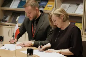 Para podpisująca dokumenty, z polską flagą obok. W tle półki z książkami, w tym te dotyczące slawistyki.